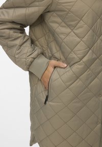 Veste d'extérieur matelassée en beige clair, avec des coutures diagonales, des poignets côtelés et une poche zippée avec quincaillerie noire.