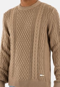 Maglione lavorato a maglia beige con motivi a treccia testurizzati, colletto e orlo a costine, con una piccola etichetta con logo sul lato inferiore anteriore.