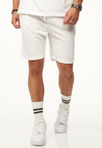 Weiße texturierte Shorts mit vertikalen Rippungen, ausgestattet mit einem elastischen Bund und einem Kordelzug. Kombiniert mit weißen Sneakern und Socken mit schwarzen Streifen.