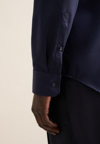 Camicia da dress blu navy che mostra il polsino con chiusura a bottone, realizzata in tessuto liscio, con un marchio discreto sulla manica.