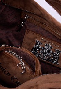 Interno di una borsa a tracolla in suede marrone con tasche con zip, caratterizzato da un'accenno di catena argentata, logo Juicy Couture e una fodera monogrammata.