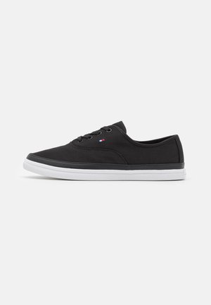 Black canvas sneakers with a low-top design, white rubber sole, and subtle branding detail on the side. Minimalistic and streamlined shape.