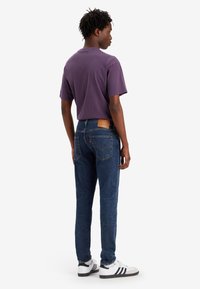 Camiseta de manga corta morada, vaqueros azules con detalles en el bolsillo trasero, y zapatillas blancas con rayas negras sobre un fondo blanco.