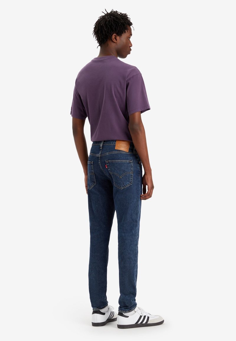 Lila Kurzarm-T-Shirt, blaue Jeans mit Rückentaschen-Detail, und weiße Sneaker mit schwarzen Streifen auf weißem Hintergrund.