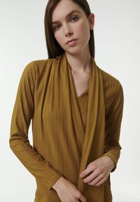 Top de manga larga en color mostaza con escote drapeado y textura acanalada, con un ajuste relajado y sutiles detalles de costura.