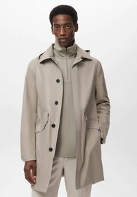 Mand iført en beige trench coat over en lys grå zip-up sweater og matchende lys beige bukser, stående mod en hvid baggrund.