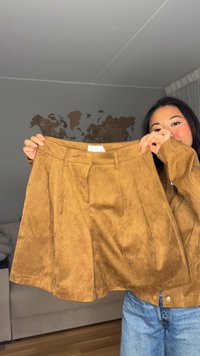 Kort brun skjørt i imitation semsket skinn med folder, med midjebånd og sidelommer. Jevn tekstur og ensartet farge.