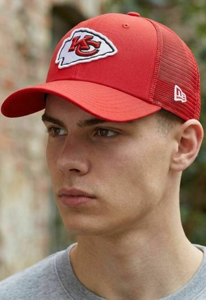 Junger Mann, der eine rote Kansas City Chiefs Baseballkappe mit Netzrückseite und ein graues Hemd trägt, schaut im Freien zur Seite.