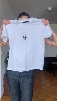 Άνθρωπος που κρατάει ένα λευκό μπλουζάκι Karl Lagerfeld με γραφικό αποτύπωμα μιας γάτας που φοράει γυαλιά ηλίου στο κέντρο της μπροστινής πλευράς.