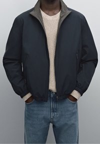 Navy bomber jacket with a khaki collar, textured knit sweater underneath, and blue straight-leg jeans. Features elastic cuffs and two pockets.