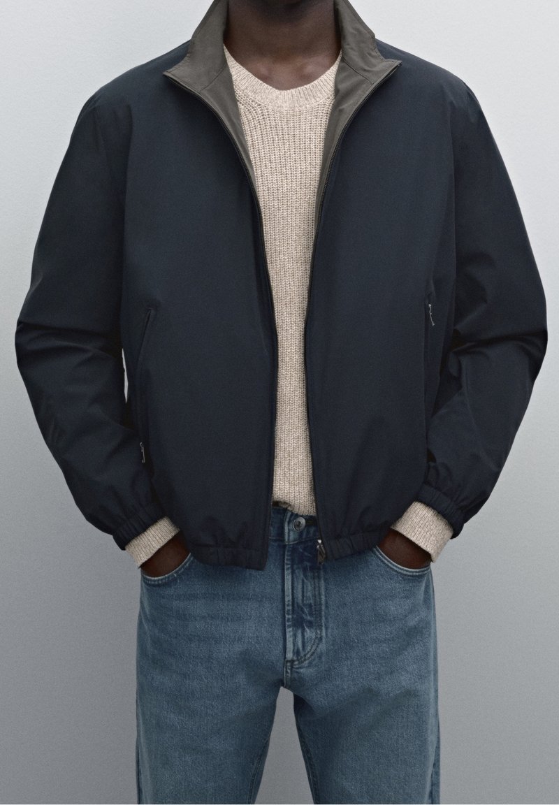 Navy bomber jacket with a khaki collar, textured knit sweater underneath, and blue straight-leg jeans. Features elastic cuffs and two pockets.