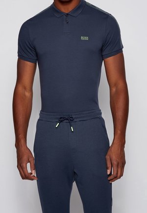 Mężczyzna noszący dopasowaną granatową koszulkę polo z logo „BOSS HUGO BOSS” na piersi oraz dopasowane granatowe spodnie ze sznurkiem.