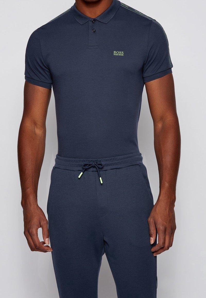 Homme portant un polo bleu marine ajusté avec le logo « BOSS HUGO BOSS » sur la poitrine et un pantalon à cordon bleu marine assorti.