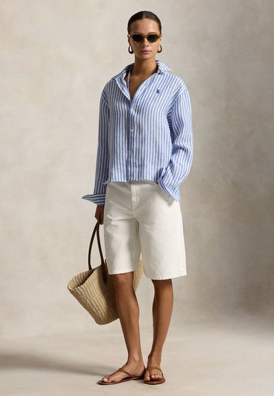 Kvinne iført blå og hvit stripete skjorte med knapper, hvite shorts til knærne, brune sandaler, solbriller, ringøreringer, holder en vevd tote-veske.