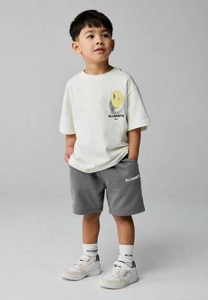 Junger Junge steht mit den Händen in den Taschen, trägt ein weißes T-Shirt mit Smiley-Gesicht, graue Shorts, weiße Socken und weiße Turnschuhe vor einem grauen Hintergrund.