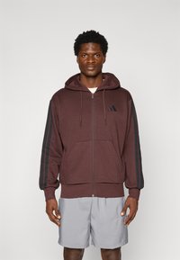 Bordeauxkleurige ritssluiting hoodie met een capuchon, zwarte strepen op de mouwen, kangoeroezakken en een klein logo op de borst. Draagt over grijze shorts.