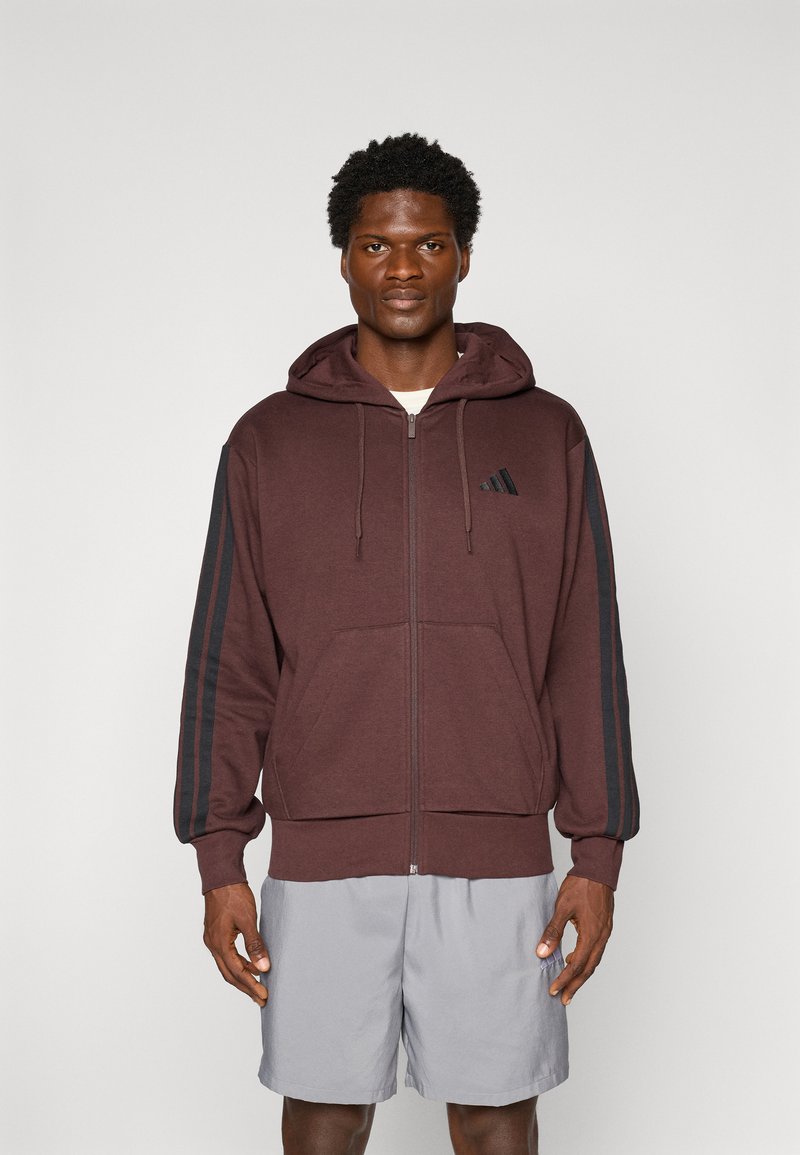 Bordeauxkleurige ritssluiting hoodie met een capuchon, zwarte strepen op de mouwen, kangoeroezakken en een klein logo op de borst. Draagt over grijze shorts.
