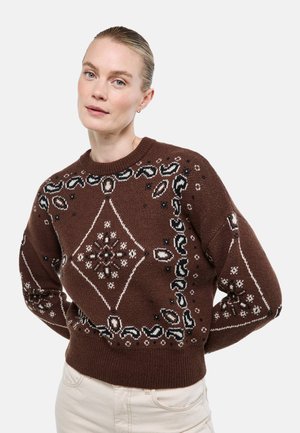 Femme aux cheveux blonds portant un pull marron en tricot avec un motif central en losange et floral, debout les mains dans le dos.