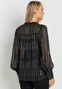 Blusa nera trasparente con maniche lunghe a sbuffo. Presenta decorazioni verticali argento e un collo rotondo. Tessuto morbido, design fluido.