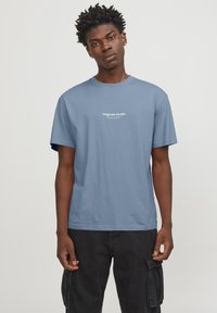 Camiseta de algodón de color azul claro con mangas cortas, que presenta un logotipo de texto blanco "Originals Studio" en el pecho. Diseño simple y de corte relajado.