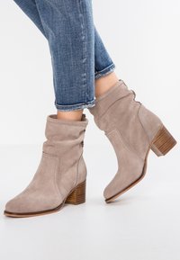 Bottes bottines en cuir suédé beige avec un design froncé, un talon empilé bas et des coutures décoratives le long des coutures. Portées avec un jean bleu retroussé.