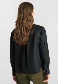 Black long-sleeve shirt made of smooth fabric, featuring a back pleat, buttoned cuffs, and a relaxed fit, paired with olive green pants.