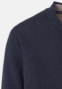 Närbild av en marinblå, texturerad tröja med axel och krage som har en beige innerfoder och tunna randiga detaljer.