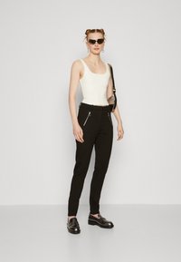 Bela skrajšana majica brez rokavov, črne slim-fit hlače z srebrnimi žepi na zadrgo, črne loafer čevlje in temne sončne očala, postavljeni na enobarvni ozadju.