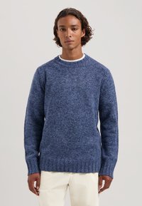 Gebreide blauwe trui met een gestructureerde afwerking, ronde halslijn en ribgebreide manchetten en zoom. Gepaard met lichtgekleurde broeken.