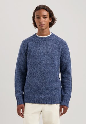 Blauer Strickpullover mit strukturiertem Finish, Rundhalsausschnitt und gerippten Bündchen und Saum. Kombiniert mit hellfarbenen Hosen.