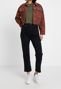 Giacca in denim marrone con tasche frontali e cuciture a contrasto, abbinata a una maglietta verde oliva, jeans neri a vita alta e sneakers bianche.