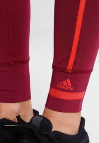 Bordové sportovní legíny s oranžovými bočními pruhy, s logem "adidas" a "Stella McCartney" na manžetě. Elastický, přiléhavý materiál.