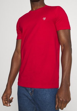 Homme portant un t-shirt rouge uni à manches courtes avec un petit logo blanc sur la poitrine gauche et un jean bleu, debout devant un fond clair.