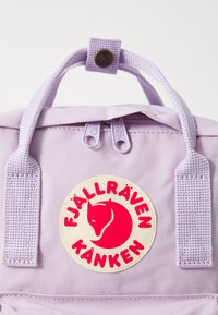 Paarse stoffen rugzak met twee bovenhandvatten, voorzien van een ronde crèmekleurige badge met een rode vos-logo en de tekst "Fjällräven Kanken."