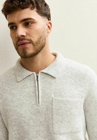 Maglione lavorato a maglia grigio chiaro con scollatura a zip, colletto piatto e taschino sul petto. Texture morbida con un motivo a coste e leggere variazioni nel colore.