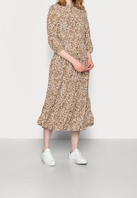 Frau in einem braun-weißen gemusterten Midi-Kleid mit Dreiviertelärmeln und weißen Turnschuhen, die auf einem grauen Boden steht.