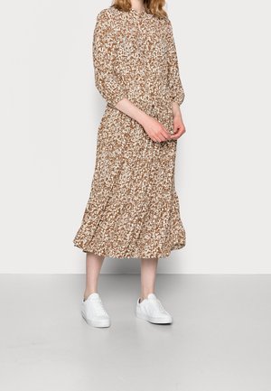 Frau in einem braun-weißen gemusterten Midi-Kleid mit Dreiviertelärmeln und weißen Turnschuhen, die auf einem grauen Boden steht.