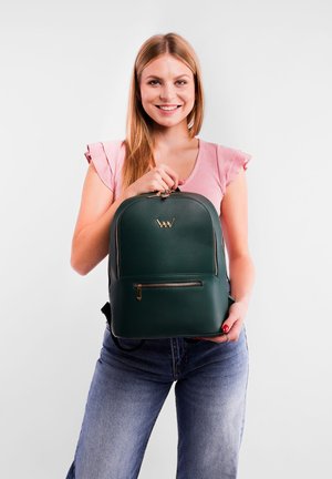 Green leather backpack with gold hardware, rounded shape, front zip pocket, and visible logo. Model holds the bag, wearing a pink top and jeans.