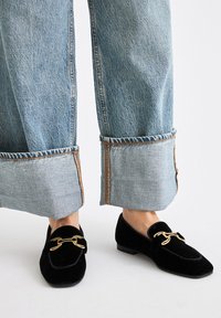 Mocasines de terciopelo negro con un detalle de cadena dorada, combinados con jeans de pierna ancha de color azul claro con dobladillos deshilachados y costuras contrastantes.