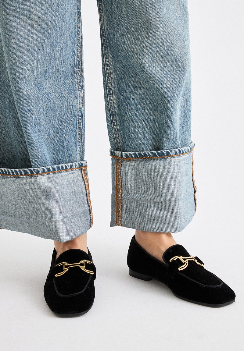 Mocasines de terciopelo negro con un detalle de cadena dorada, combinados con jeans de pierna ancha de color azul claro con dobladillos deshilachados y costuras contrastantes.