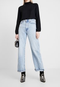 Donna che indossa una blusa nera a maniche lunghe, jeans blu chiaro a vita alta, stivaletti metallici argento alla caviglia, mentre tiene una piccola borsa nera.