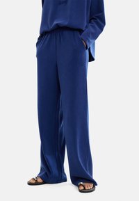 Granatowe spodnie o szerokich nogawkach z miękkiego materiału, z elastycznym paskiem i bocznymi kieszeniami, w komplecie z czarnymi sandałami.