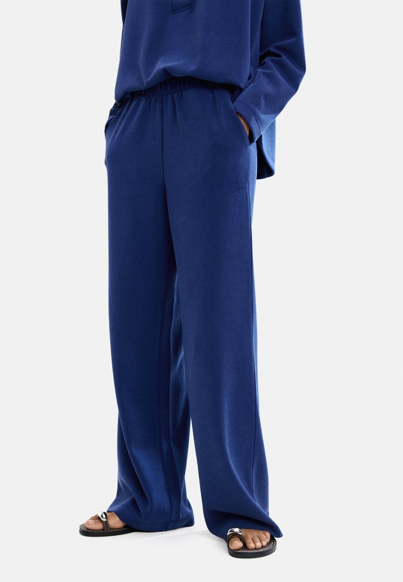 Granatowe spodnie o szerokich nogawkach z miękkiego materiału, z elastycznym paskiem i bocznymi kieszeniami, w komplecie z czarnymi sandałami.
