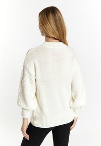 Weißer Strickpullover mit Rundhals, lockerer Passform, strukturierten Ärmeln und geripptem Saum. Getragen mit dunklen Hosen, die das Design auf der Rückseite zeigen.