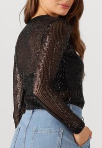 Zwart pailletten top met lange, doorschijnende mouwen en gestructureerde stof, met verticale strepen en een getailleerd ontwerp, gecombineerd met een lichtblauwe denim onderkant.