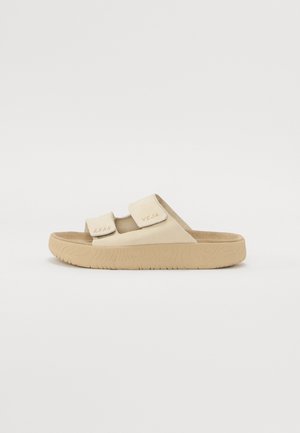 Beige slide sandals with two adjustable straps, textured upper, and a rubber sole featuring ridged detailing. Brand logo embossed on strap.