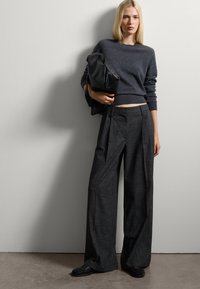 Grijze cropped sweater gecombineerd met wijde, gestructureerde donkere broek. Zwarte tas onder de arm, met één hand vastgehouden. Eenvoudige achtergrond en neutrale tinten.