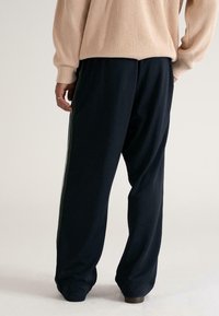 Persoon in losse zwarte broek met zijstrepen en een beige gebreide trui, staand met één hand in de zak, met de rug naar de camera.