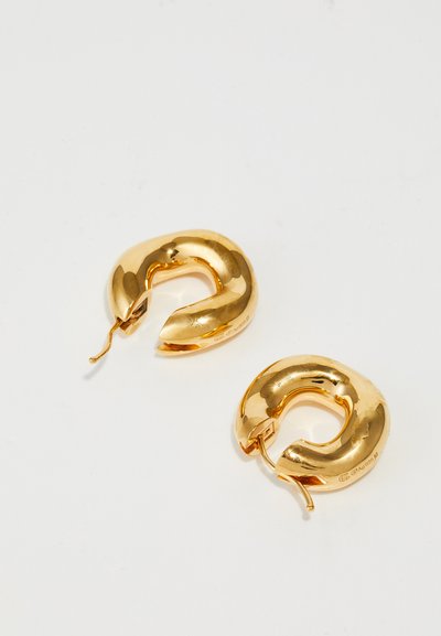 Paire de boucles d'oreilles créoles épaisses dorées et brillantes avec fermoirs ouverts reposant sur une surface blanche unie.