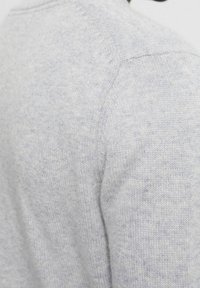 Hellgrauer Pullover mit einem strukturierten Strickmuster, der subtile blaue Flecken aufweist. Der Stoff erscheint weich und seine Form umschmeichelt den Hals und die Schultern.